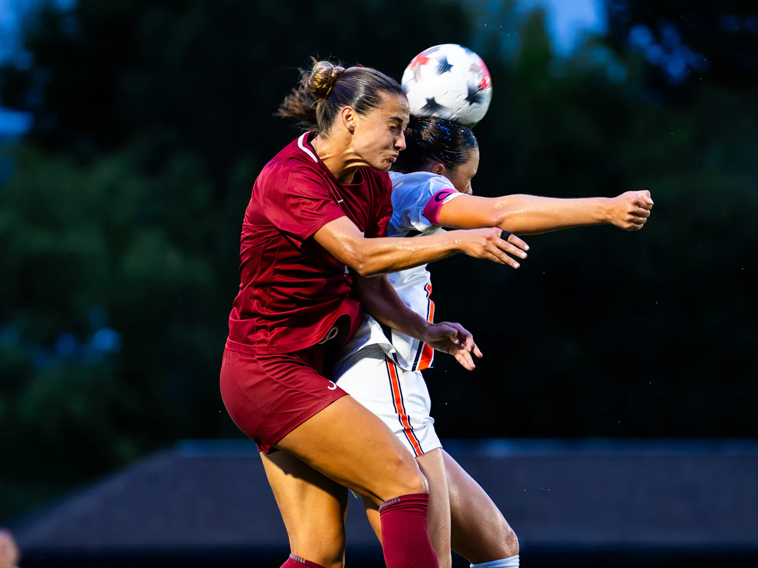 Two soccer players battle for possession of the ball during the Iron Bowl of Soccer, with an Alabama player in crimson challenging an Auburn player in white for a header.