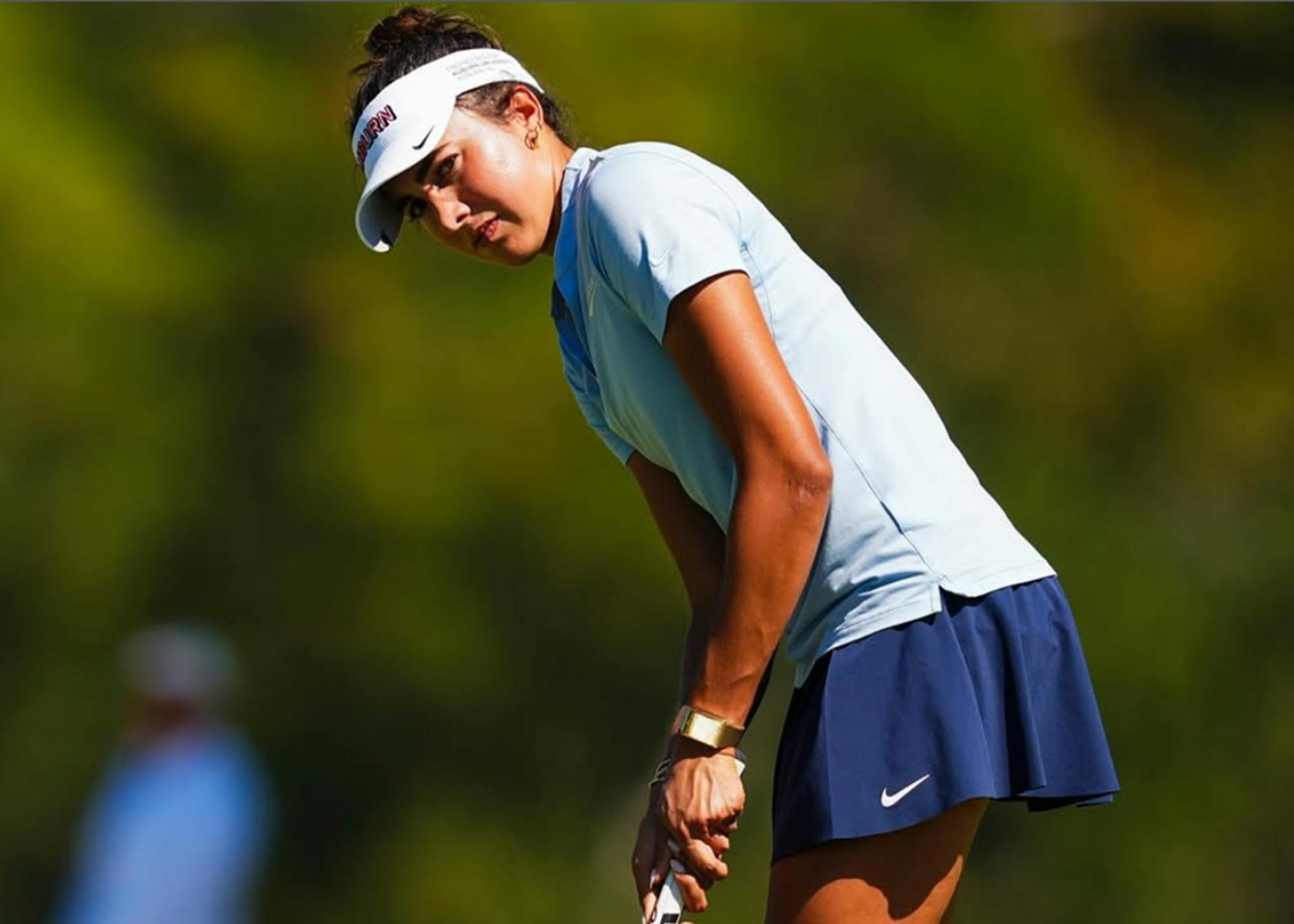 Medium close-up of Auburn women's golfer Charlotte Cantonis lining up a putt on a sunny golf course. She wears a white visor with "AUBURN" printed on the side, a light blue short-sleeved polo, and a navy blue Nike athletic skirt. Leaning forward with her hands gripped on her putter, she focuses intently on the line of her shot against a softly blurred background of lush green trees.