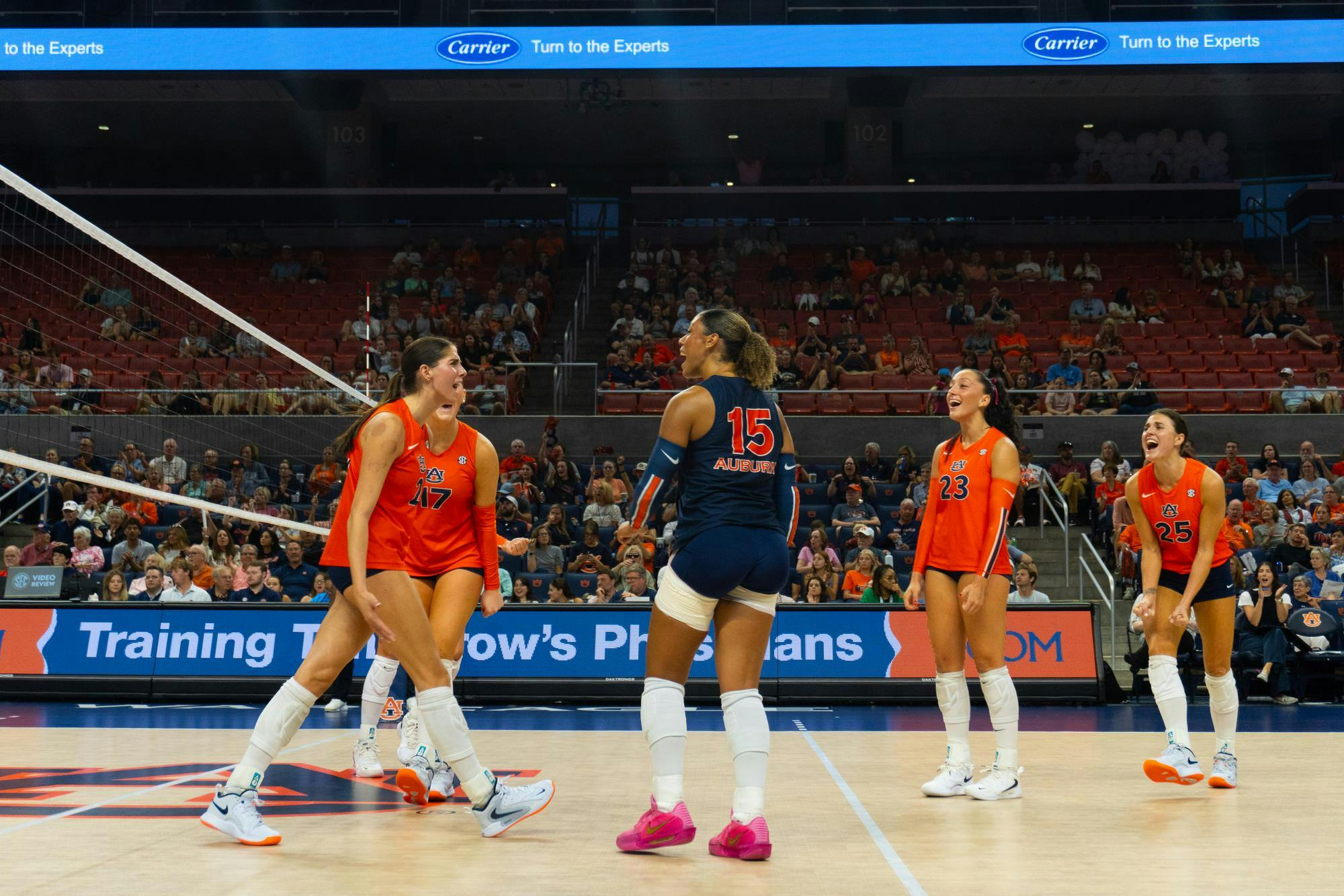 Five Auburn players celebrate on the court after winning a point, each with excited expressions. Four are wearing orange uniforms, with the player in the center wearing a navy blue libero uniform