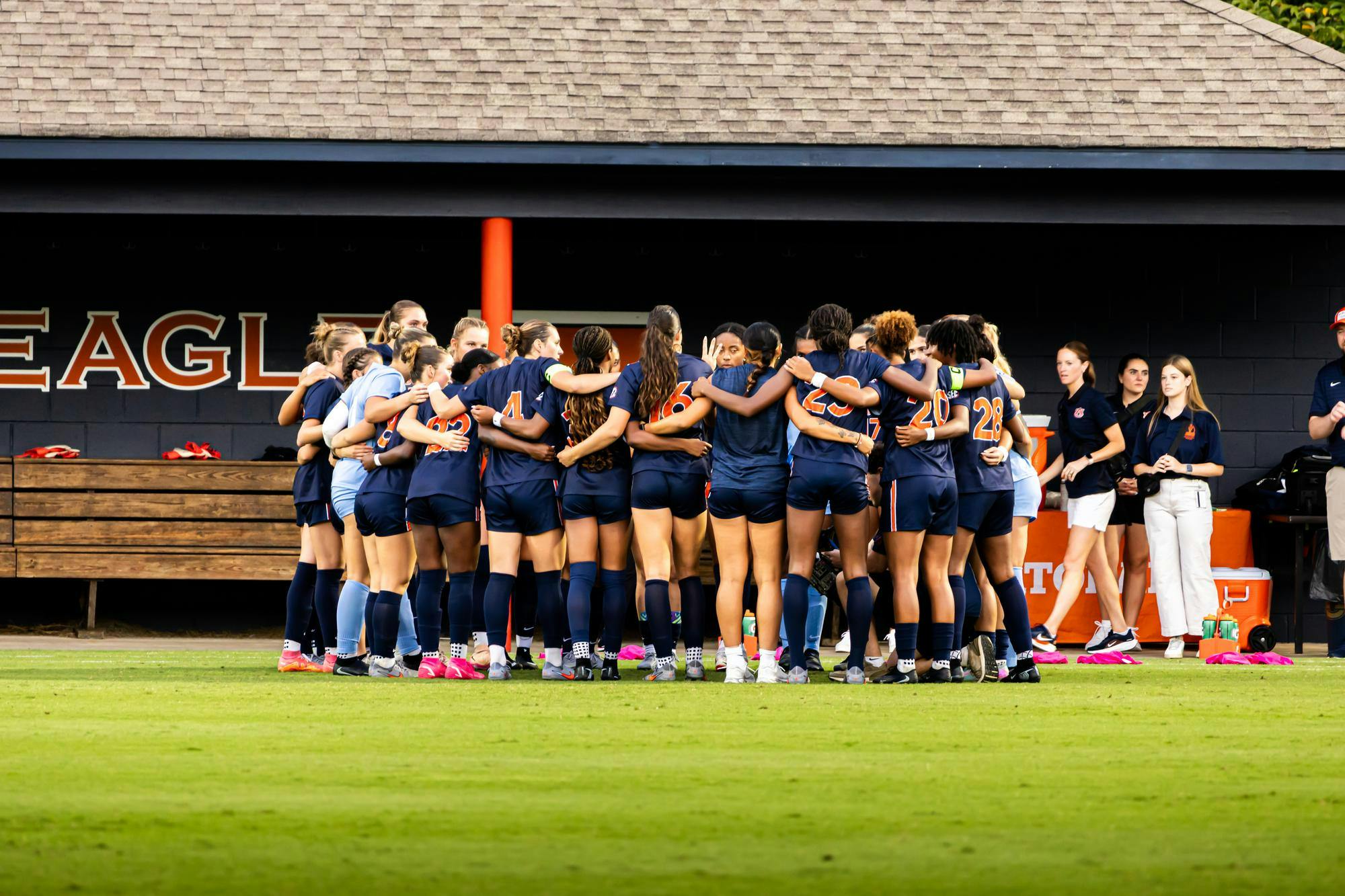 A women’s soccer team wearing navy uniforms huddles together on the field before a game, with coaches and staff standing nearby in front of the dugout.