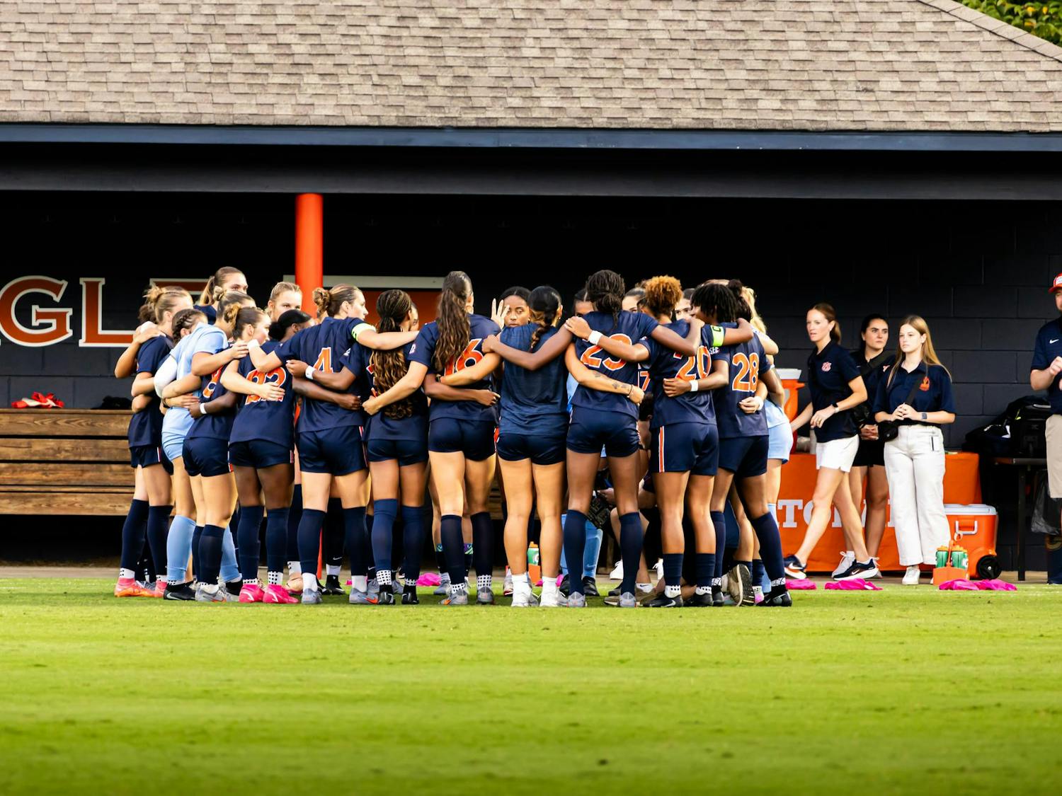 A women’s soccer team wearing navy uniforms huddles together on the field before a game, with coaches and staff standing nearby in front of the dugout.