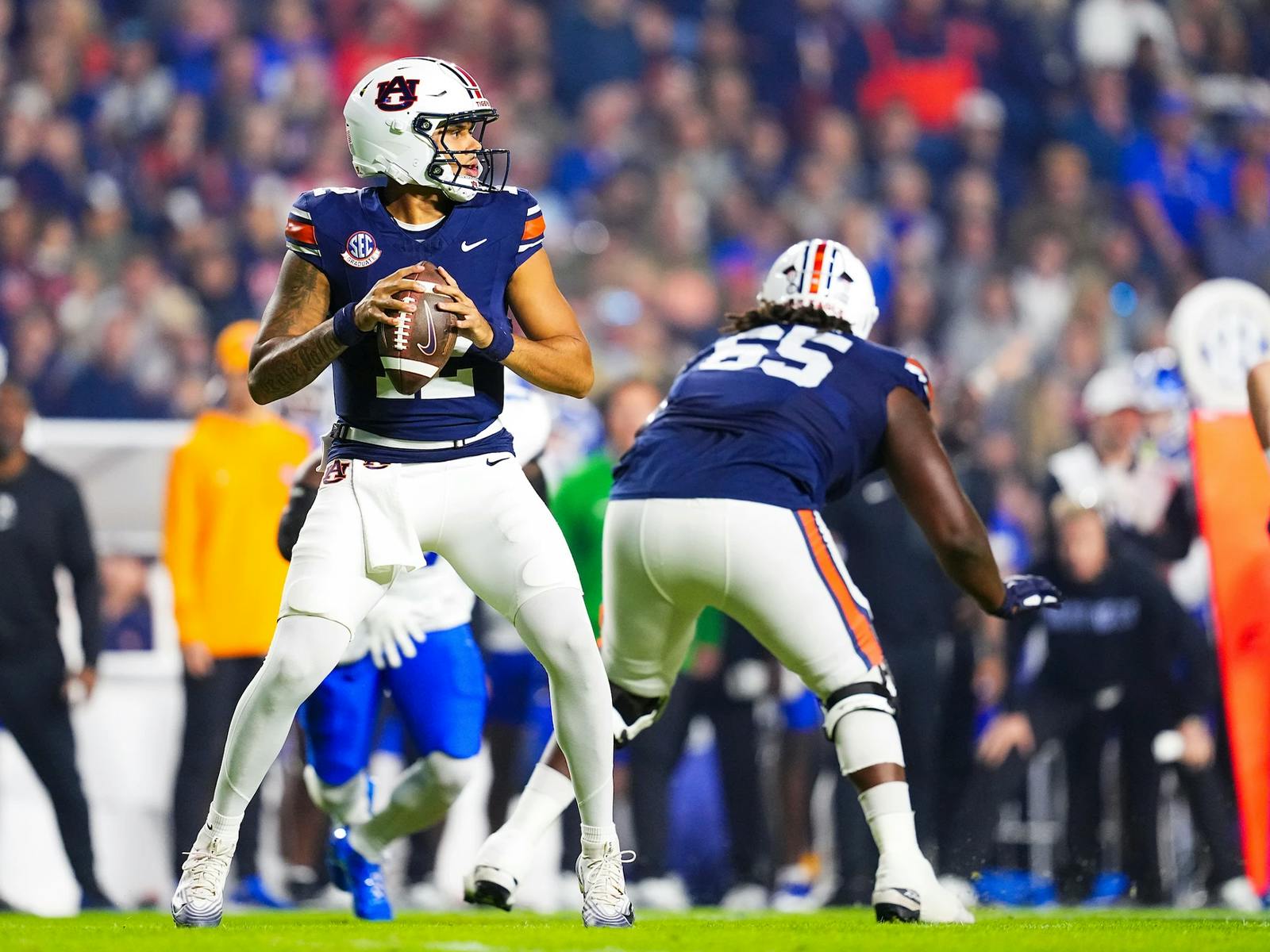 Auburn football quarterback Ashton Daniels wearing a navy blue jersey and white helmet prepares to throw the ball during a game. A teammate in a matching uniform blocks in front of him, while opposing players in blue and white approach. The play unfolds on a brightly lit field with a blurred crowd in the background.