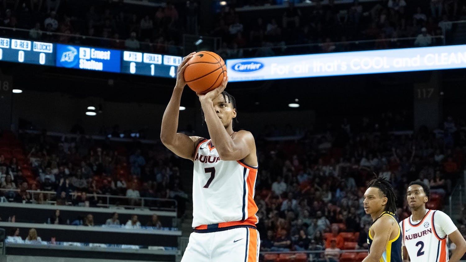 An Auburn basketball player wearing a white jersey with number 7 prepares to take a free throw during a game. He focuses intently on the basket, holding the ball in shooting position. Teammates and opposing players stand nearby, while fans fill the arena seats in the background under bright court lighting.