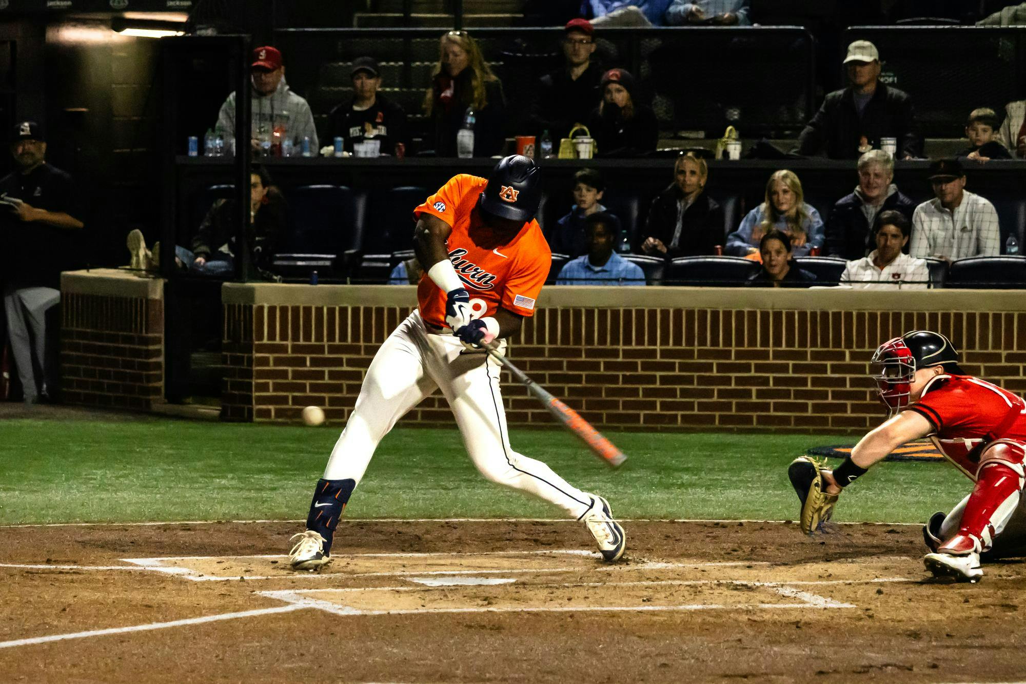 An Auburn baseball player in an orange jersey swings the bat and makes contact with a pitch at home plate during a night game. A catcher in red gear crouches to the right as the ball leaves the bat, with spectators seated behind a brick backstop in the background.