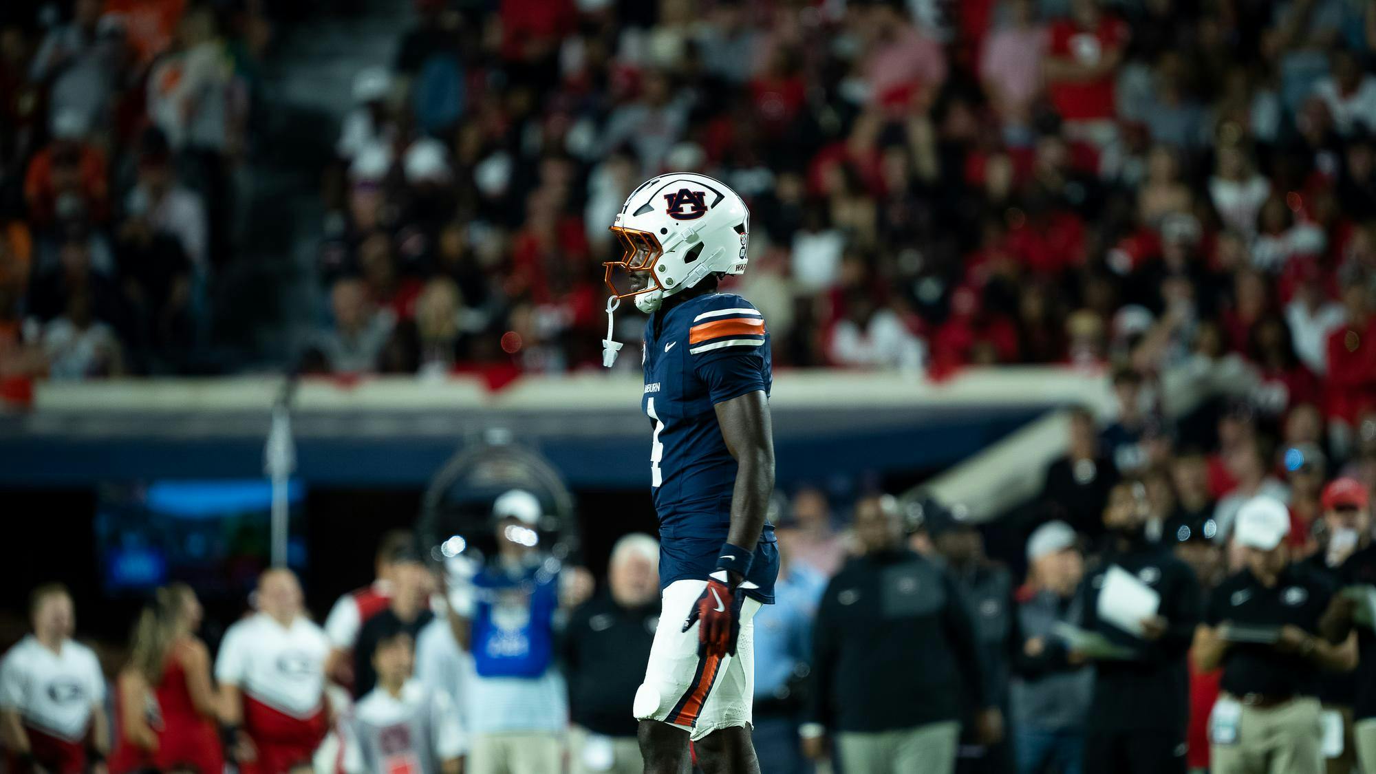 Auburn football player wearing a navy blue uniform and white helmet with the Auburn logo stands on the field during a game, with a blurred crowd and sideline staff visible in the background.