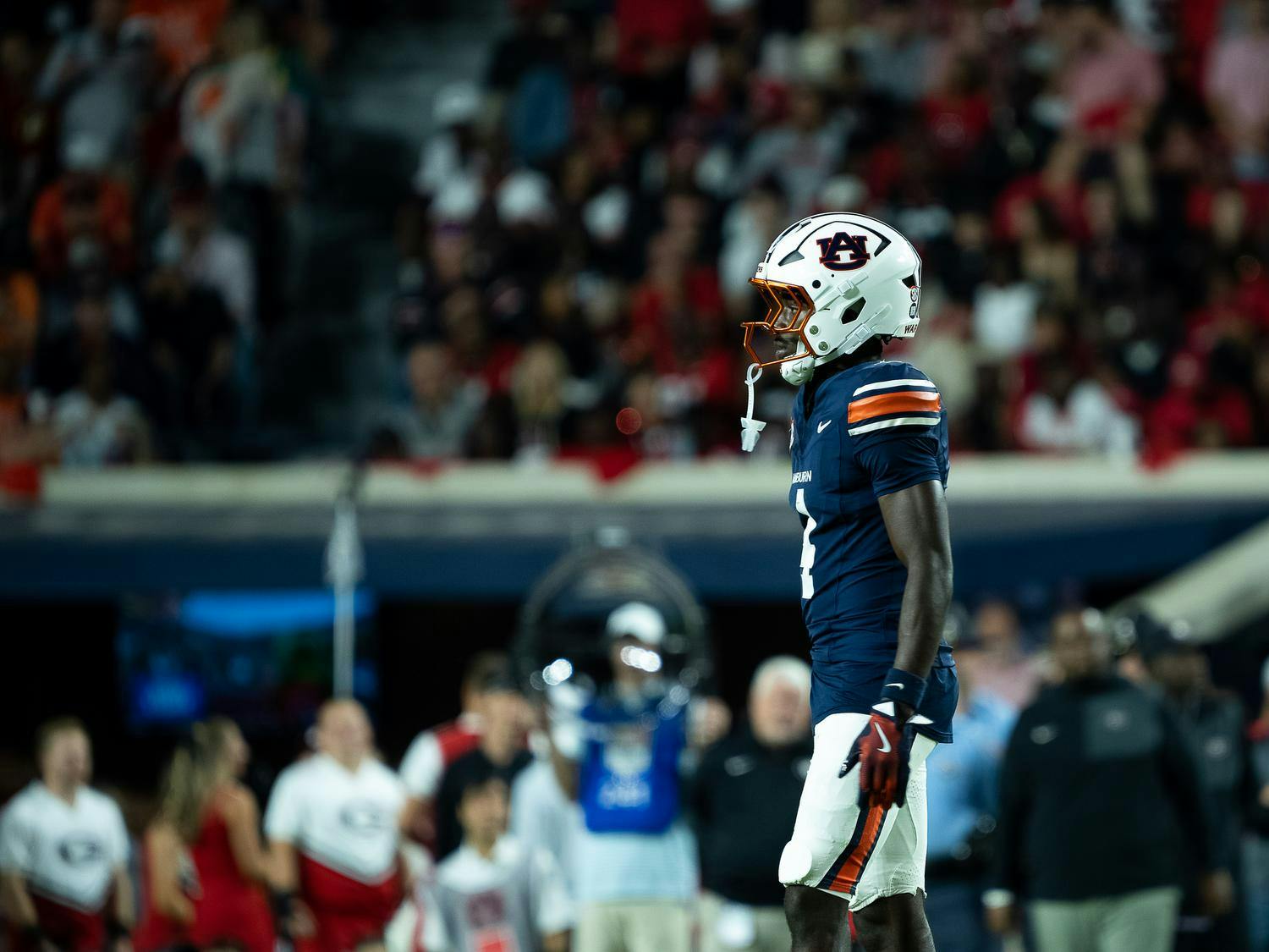 Auburn football player wearing a navy blue uniform and white helmet with the Auburn logo stands on the field during a game, with a blurred crowd and sideline staff visible in the background.