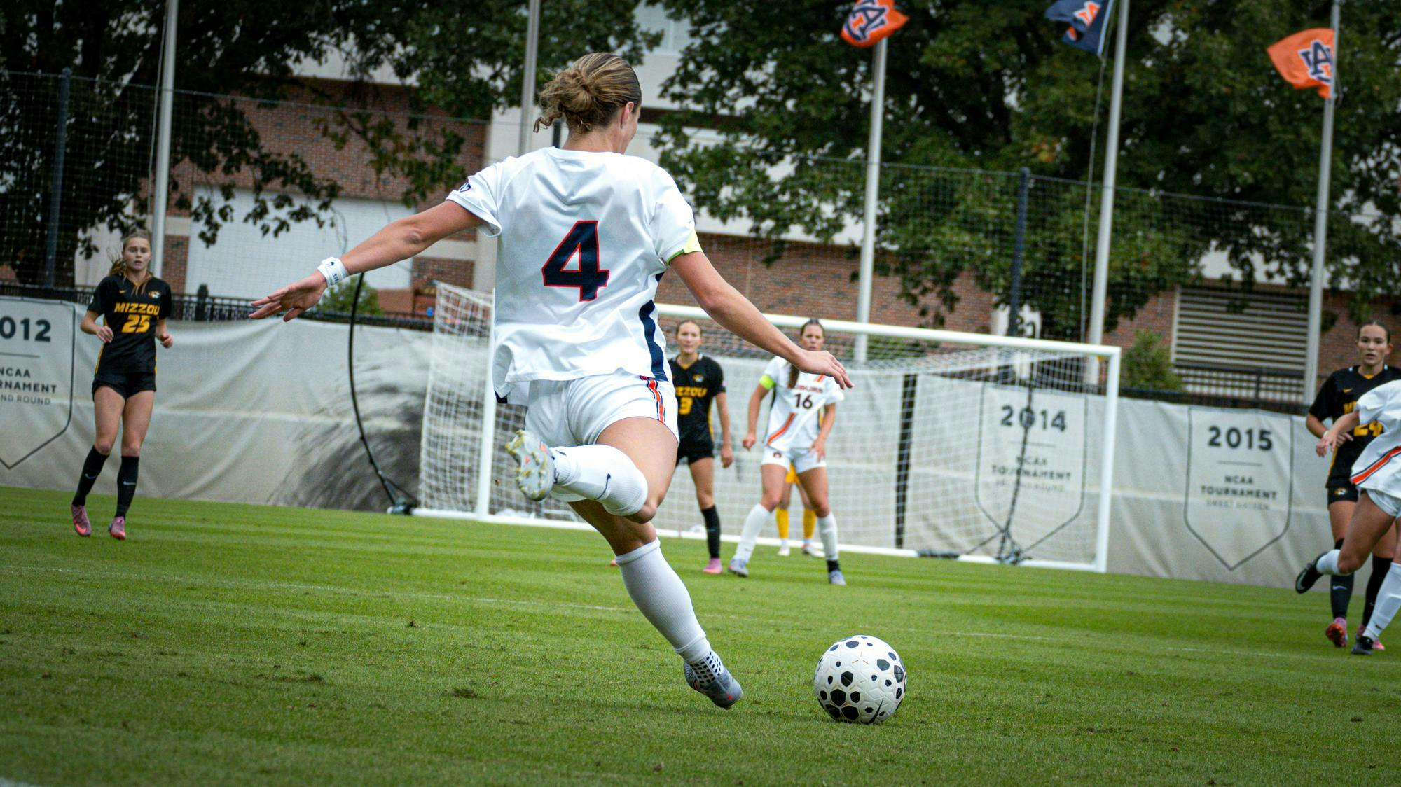Auburn soccer player wearing a white No. 4 jersey kicks the ball toward the goal during a match against Missouri, whose players wear black and gold uniforms. Several players and Auburn banners are visible in the background, along with NCAA tournament year signs hanging behind the goal.