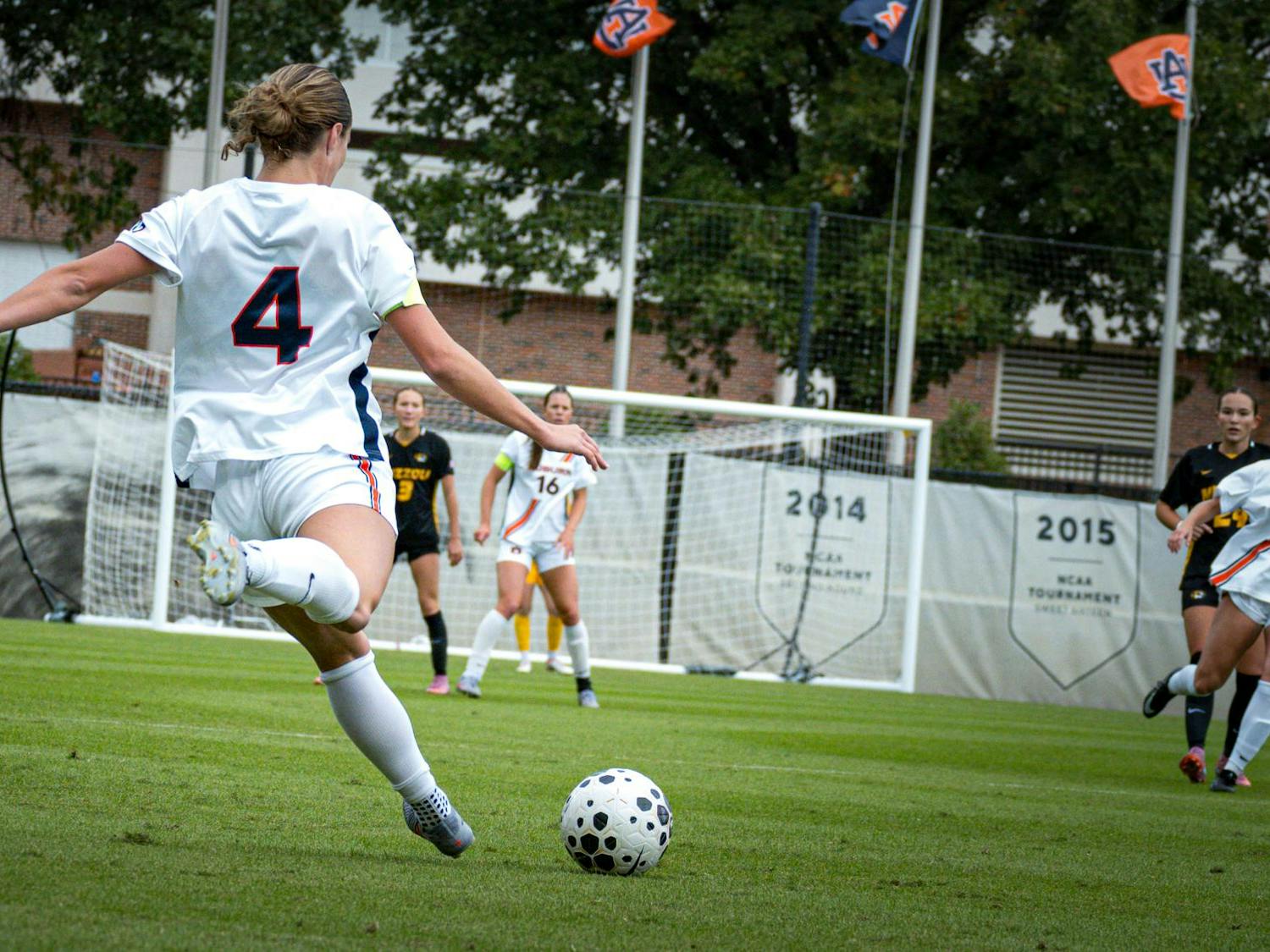 Auburn soccer player wearing a white No. 4 jersey kicks the ball toward the goal during a match against Missouri, whose players wear black and gold uniforms. Several players and Auburn banners are visible in the background, along with NCAA tournament year signs hanging behind the goal.