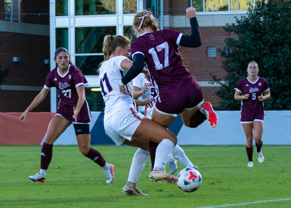 A high-action shot from a women's college soccer match between Auburn and Mississippi State. An Auburn player in a white uniform fights for possession of the ball against a Mississippi State defender wearing a maroon jersey with the number 17. The defender is airborne, leaning into the challenge as they both contest the ball on the green grass field. In the background, two other Mississippi State players in maroon jerseys track the play closely near the sideline.