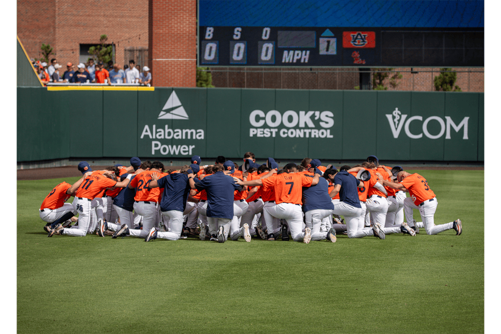 Players and coaches from the Auburn baseball team kneel shoulder-to-shoulder in a circle on the outfield grass at Plainsman Park before the start of a Super Regional game against Ole Miss. Fans and the outfield scoreboard are visible in the background.
