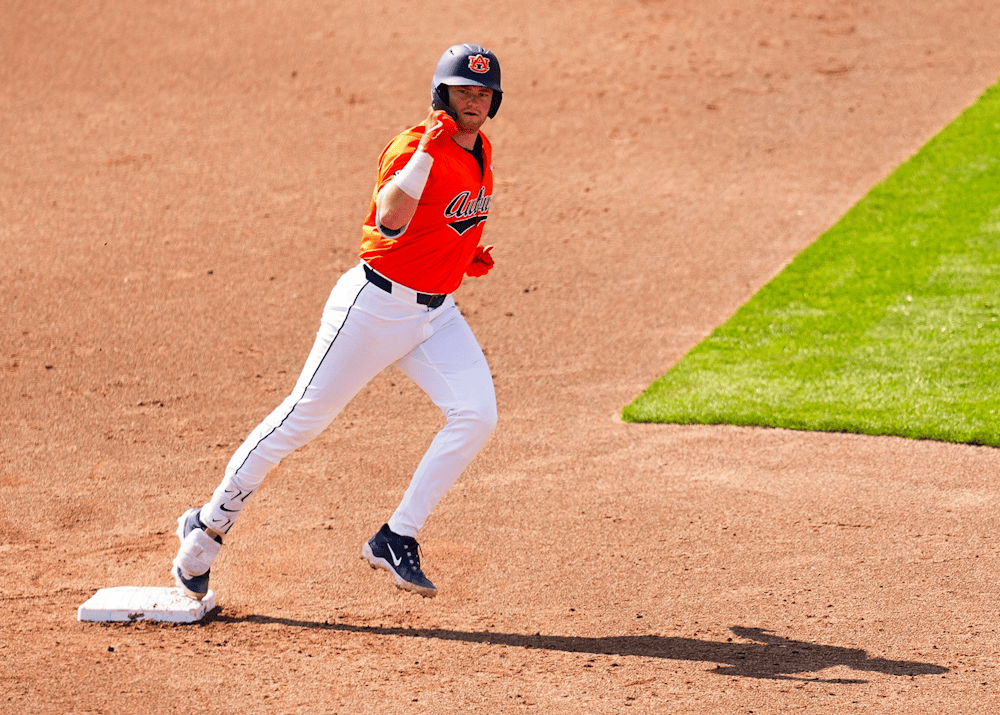 An Auburn baseball player wearing an orange jersey and gray pants rounds a base on the infield dirt, lifting one hand as he runs. He wears a navy helmet with an Auburn logo, and bright green outfield grass contrasts with the brown infield behind him.