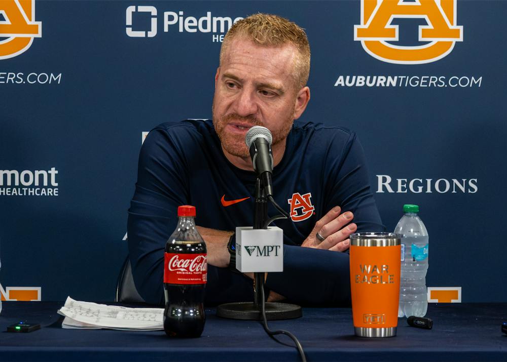 Auburn head coach Alex Golesh speaks into a microphone during a press conference at a table with water bottles and a drink, with Auburn logos and sponsor signage displayed on the backdrop.