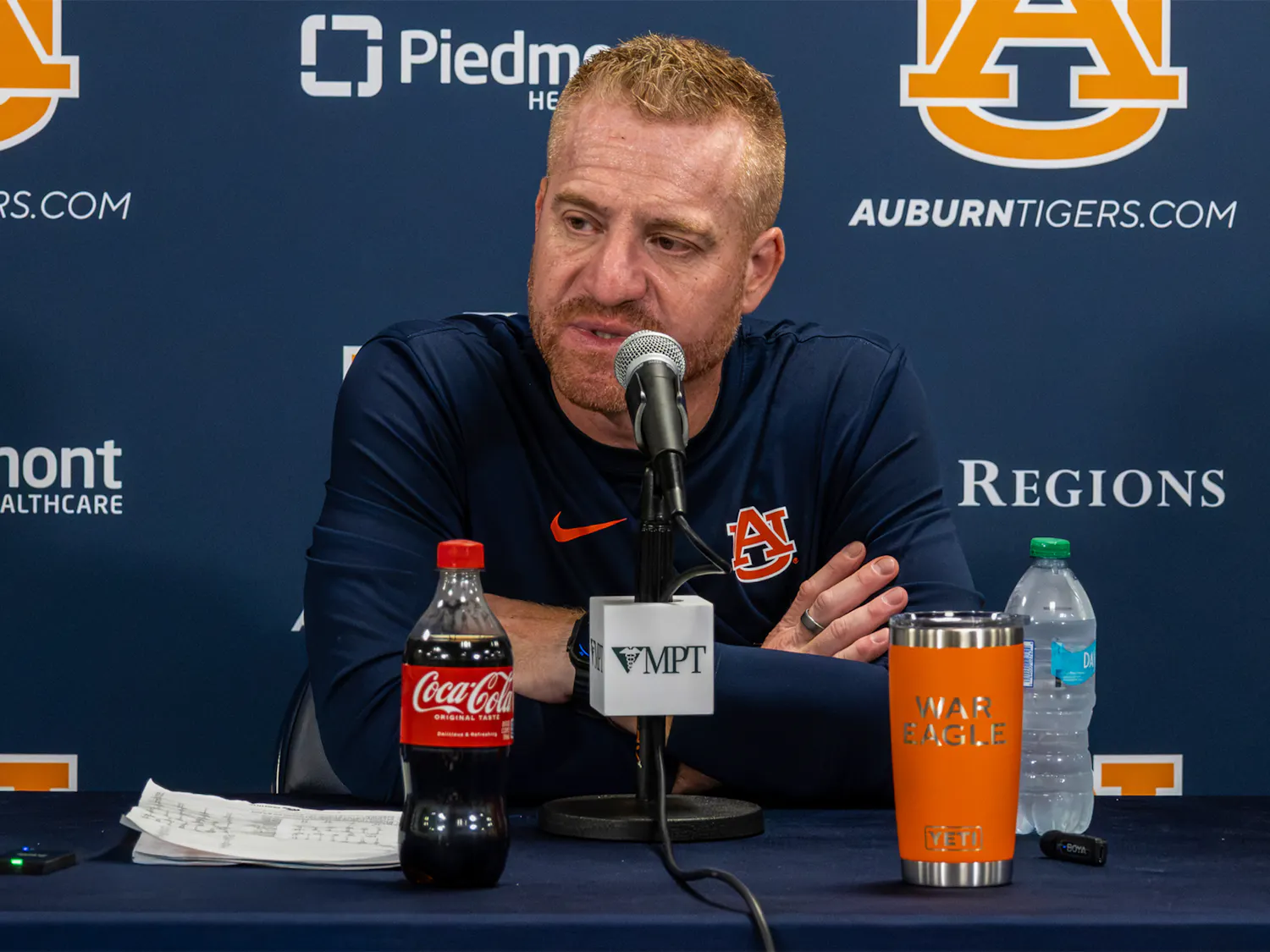 Auburn head coach Alex Golesh speaks into a microphone during a press conference at a table with water bottles and a drink, with Auburn logos and sponsor signage displayed on the backdrop.