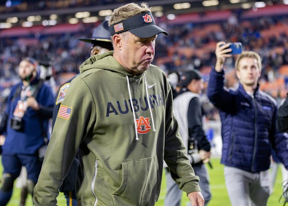 Hugh Freeze wearing an olive-green Auburn hoodie and a black visor walks off the field with a serious expression. People and media personnel surround him, with one person taking a photo or video on a phone. The background shows a stadium with blurred spectators and bright lights.
