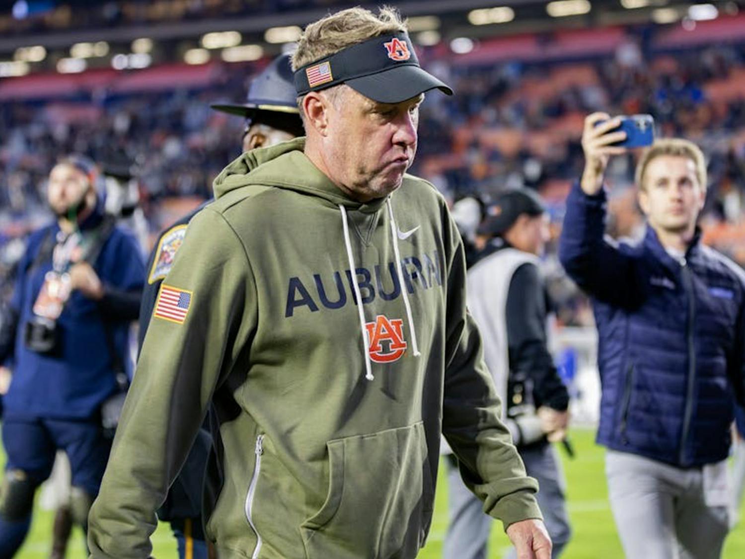 Hugh Freeze wearing an olive-green Auburn hoodie and a black visor walks off the field with a serious expression. People and media personnel surround him, with one person taking a photo or video on a phone. The background shows a stadium with blurred spectators and bright lights.