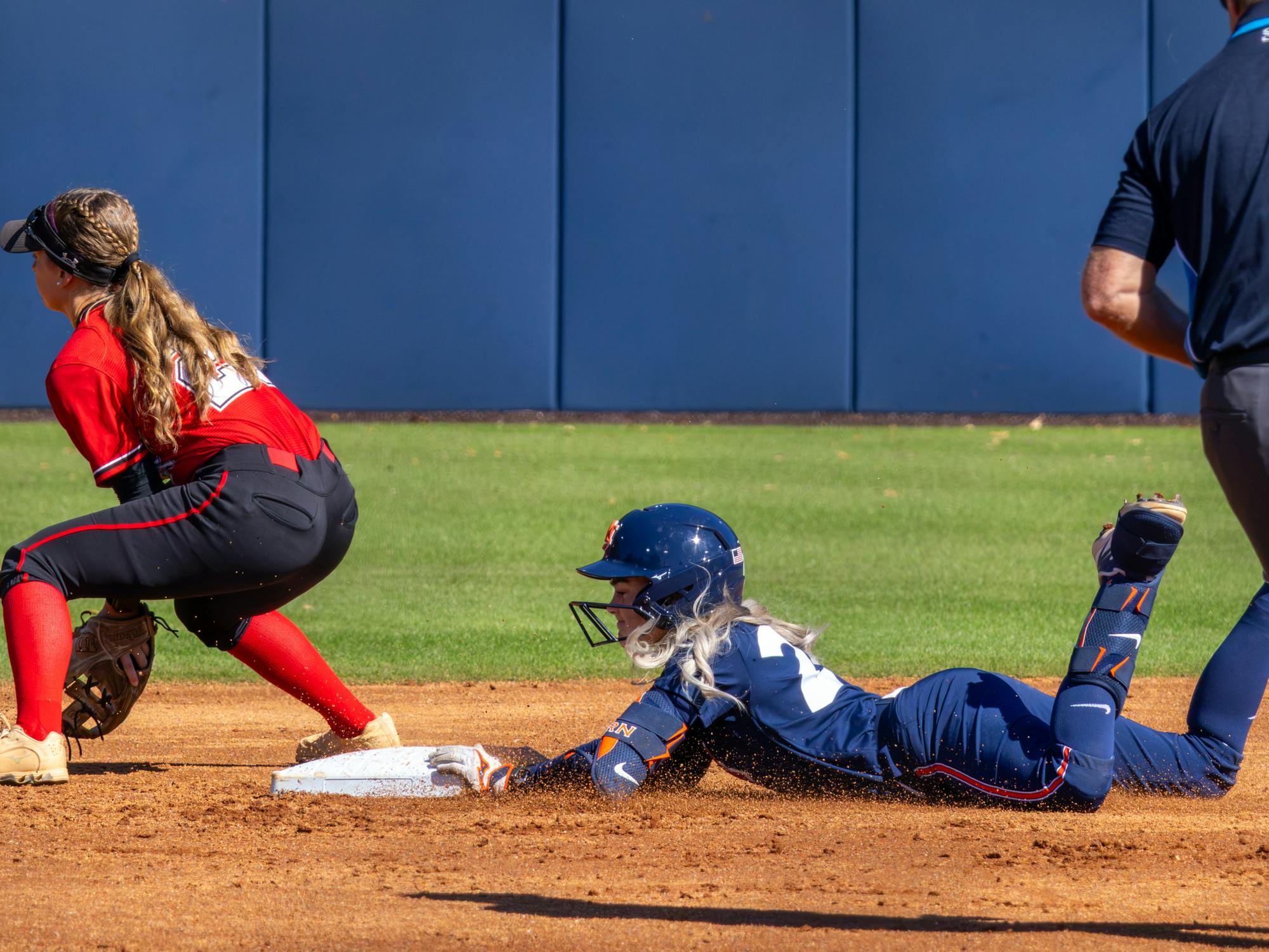 An Auburn softball player in a navy blue uniform with an elbow guard slides headfirst along the dirt toward a base, extending her gloved left hand to touch the white canvas. A Gadsden State Cardinals fielder in a red jersey, dark pants, and red socks crouches in position near the bag as an umpire stands to the right.