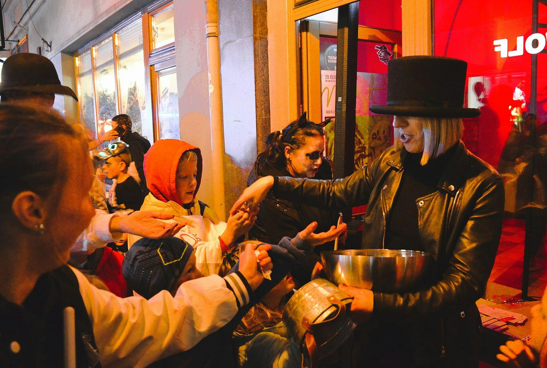 Children in Halloween costumes gather around a woman in a top hat as she hands out candy on a brightly lit street 