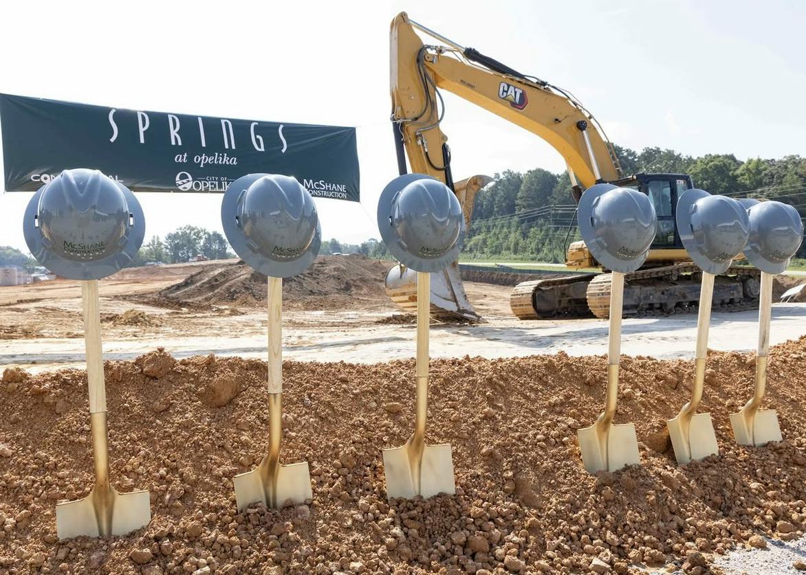 Six ceremonial shovels topped with silver hard hats stand in freshly turned soil at the Springs at Opelika groundbreaking, with a construction excavator and project banner in the background.
