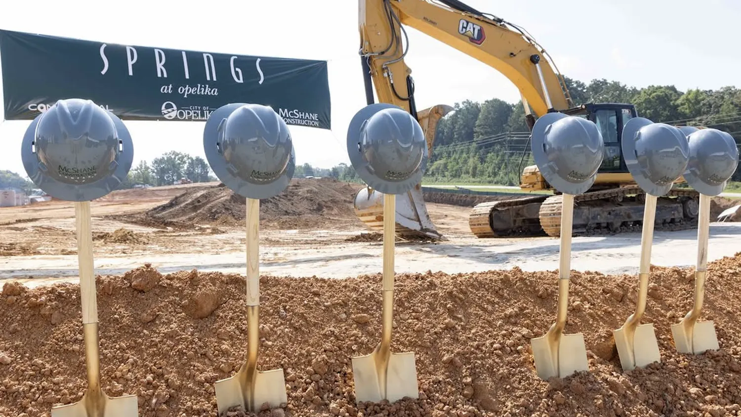Six ceremonial shovels topped with silver hard hats stand in freshly turned soil at the Springs at Opelika groundbreaking, with a construction excavator and project banner in the background.