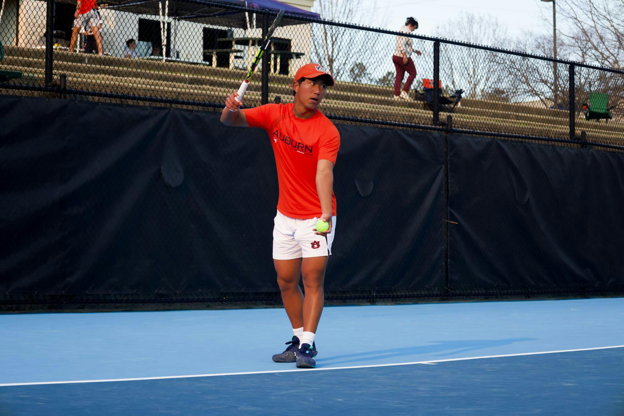 Auburn men’s tennis player in an orange shirt and white shorts prepares to serve during a match on an outdoor court.