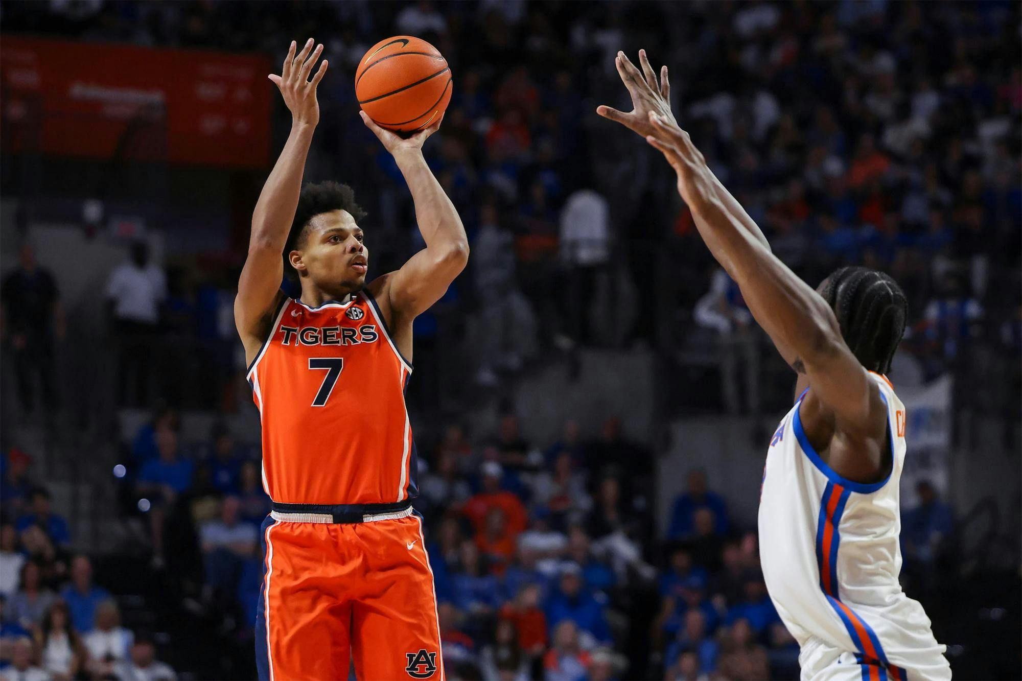 Auburn men’s basketball player in an orange No. 7 jersey jumps and shoots a jump shot over a Florida defender with arms raised in a packed indoor arena, with the crowd blurred in the background.
