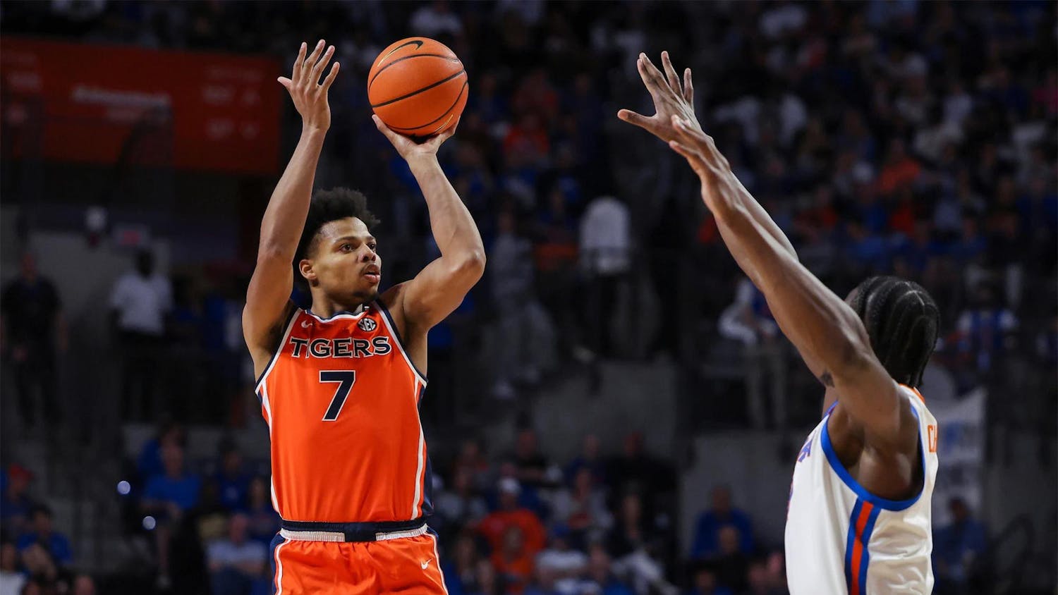 Auburn men’s basketball player in an orange No. 7 jersey jumps and shoots a jump shot over a Florida defender with arms raised in a packed indoor arena, with the crowd blurred in the background.