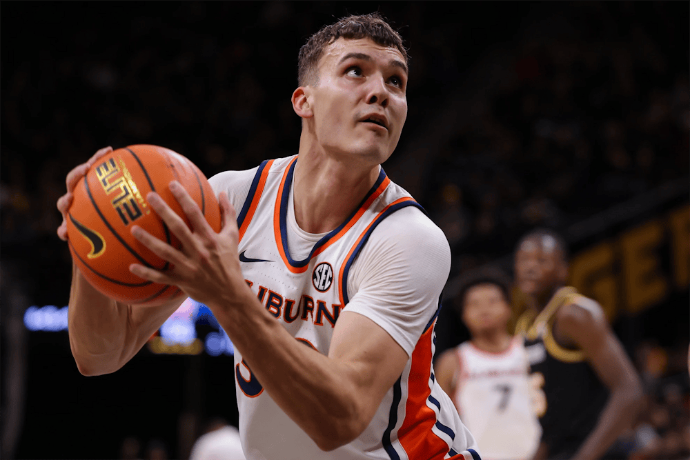 Auburn men’s basketball player in a white jersey grips the ball with both hands and looks toward the basket while driving inside, with blurred defenders and arena crowd in the background.