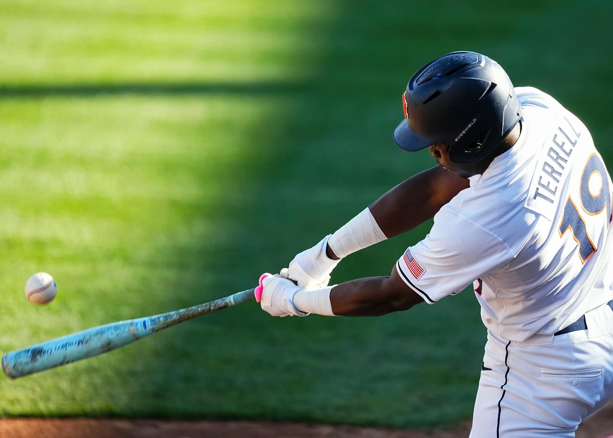 A baseball player in a white uniform with “TERRELL” and the number 19 on the back swings a bat, making contact with a pitch as the ball flies off to the left. He wears a dark helmet and batting gloves, and the action is captured mid-swing against a blurred green outfield background.