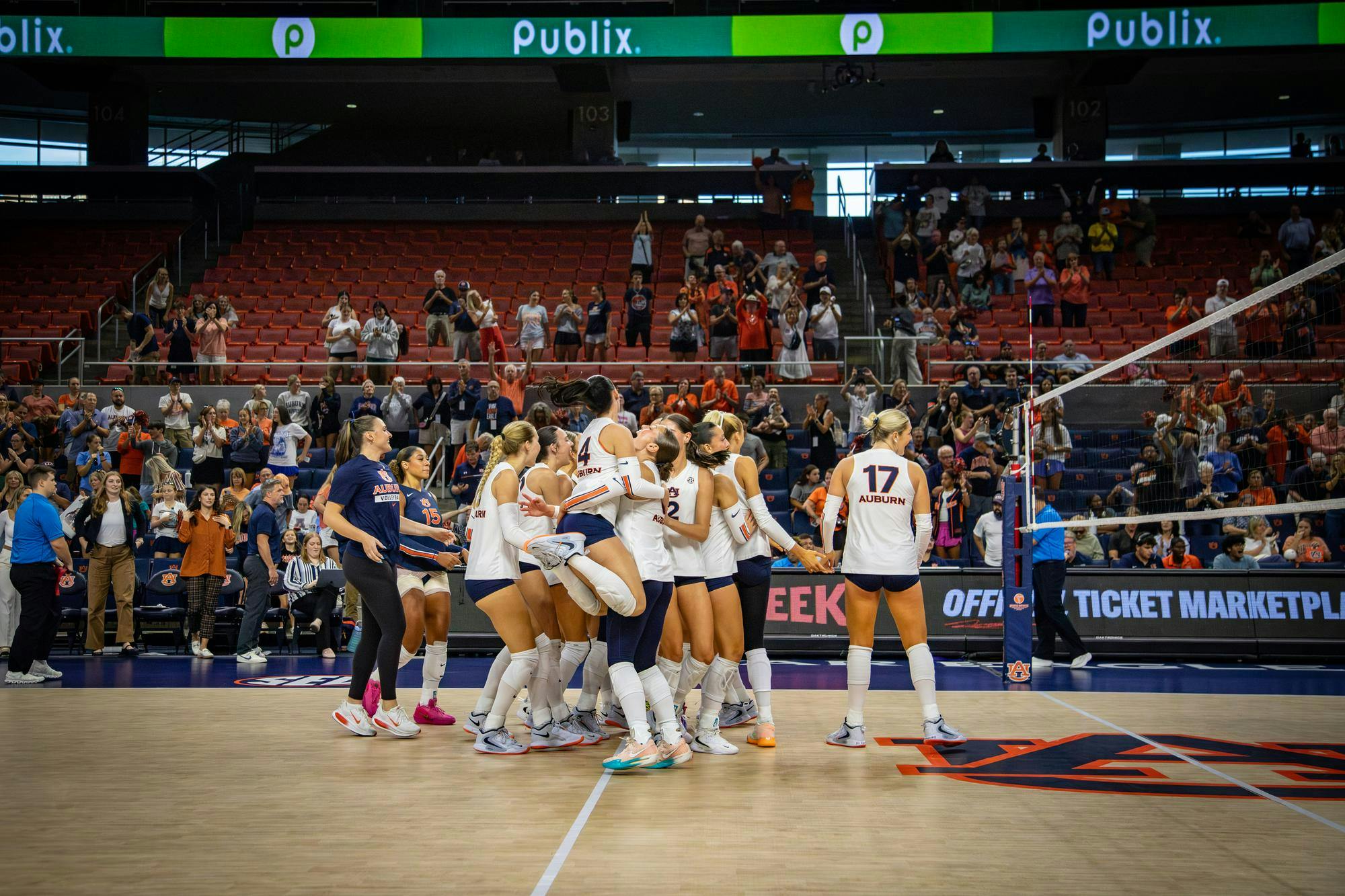 Auburn women’s volleyball players celebrate on the court in front of cheering fans after a victory, with teammates hugging and jumping together near the net inside an arena.