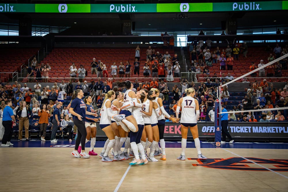 Auburn women’s volleyball players celebrate on the court in front of cheering fans after a victory, with teammates hugging and jumping together near the net inside an arena.