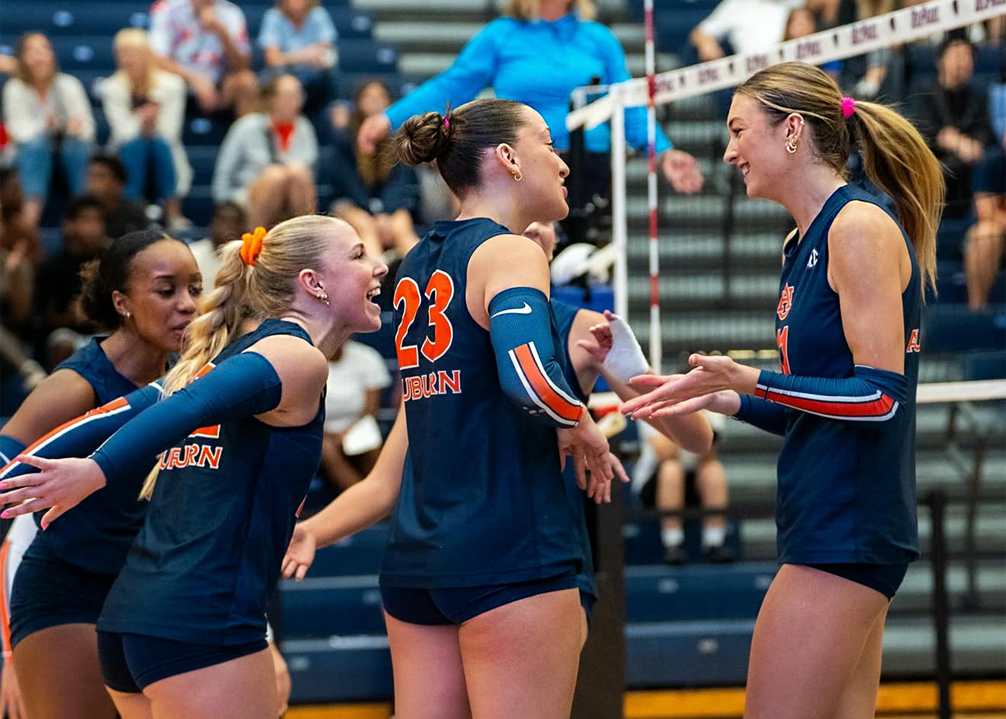 Auburn volleyball players celebrate together near the net during a match, with four players in navy uniforms visible in the foreground.