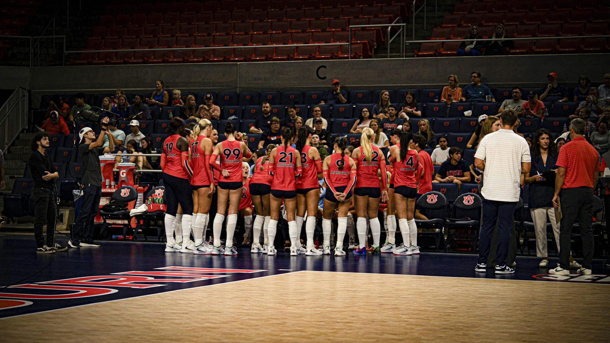 A women’s volleyball team wearing orange jerseys gathers in a huddle near the bench before a match, with coaches and staff standing nearby and fans watching from the stands in the background.