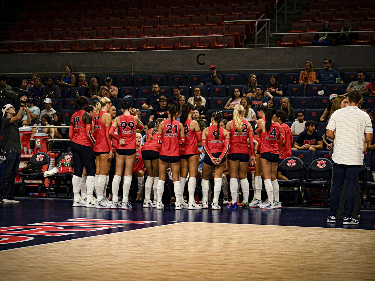 A women’s volleyball team wearing orange jerseys gathers in a huddle near the bench before a match, with coaches and staff standing nearby and fans watching from the stands in the background.