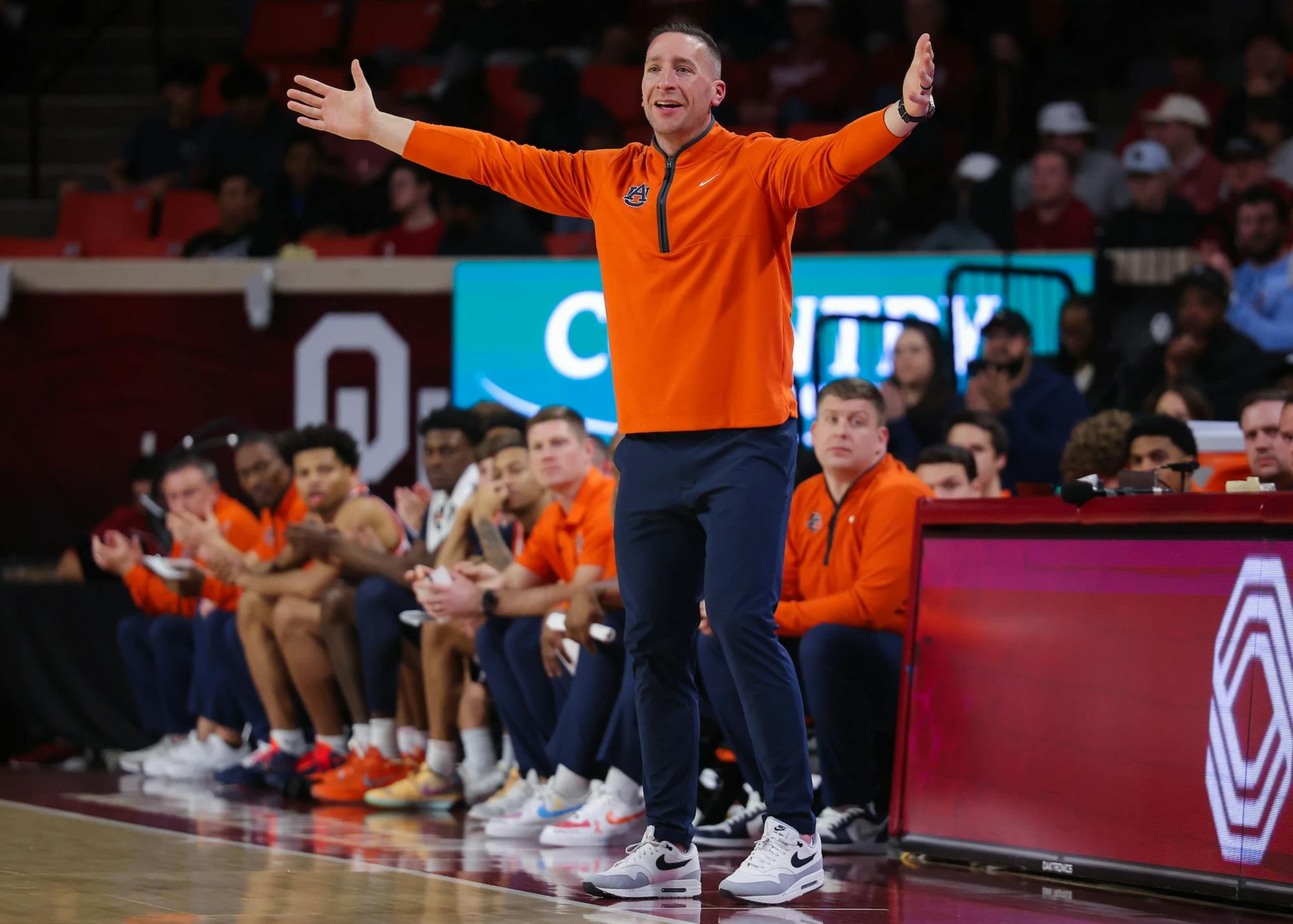 A college basketball coach wearing an orange quarter-zip pullover and navy pants stands on the sideline with his arms stretched wide, reacting to the action on the court. Players and staff in matching orange sit on the bench behind him, while an arena backdrop featuring a large “OU” logo and spectators fills the background.