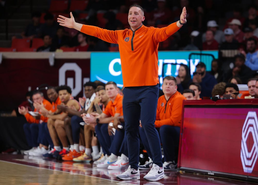 A college basketball coach wearing an orange quarter-zip pullover and navy pants stands on the sideline with his arms stretched wide, reacting to the action on the court. Players and staff in matching orange sit on the bench behind him, while an arena backdrop featuring a large “OU” logo and spectators fills the background.