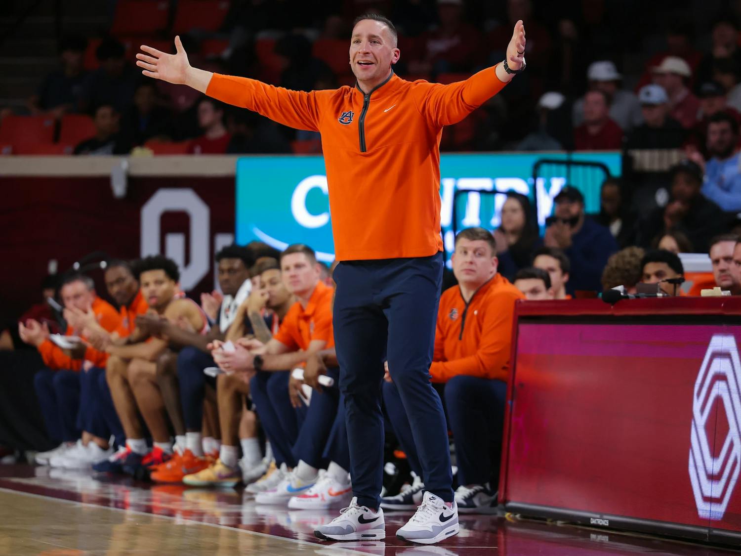 A college basketball coach wearing an orange quarter-zip pullover and navy pants stands on the sideline with his arms stretched wide, reacting to the action on the court. Players and staff in matching orange sit on the bench behind him, while an arena backdrop featuring a large “OU” logo and spectators fills the background.