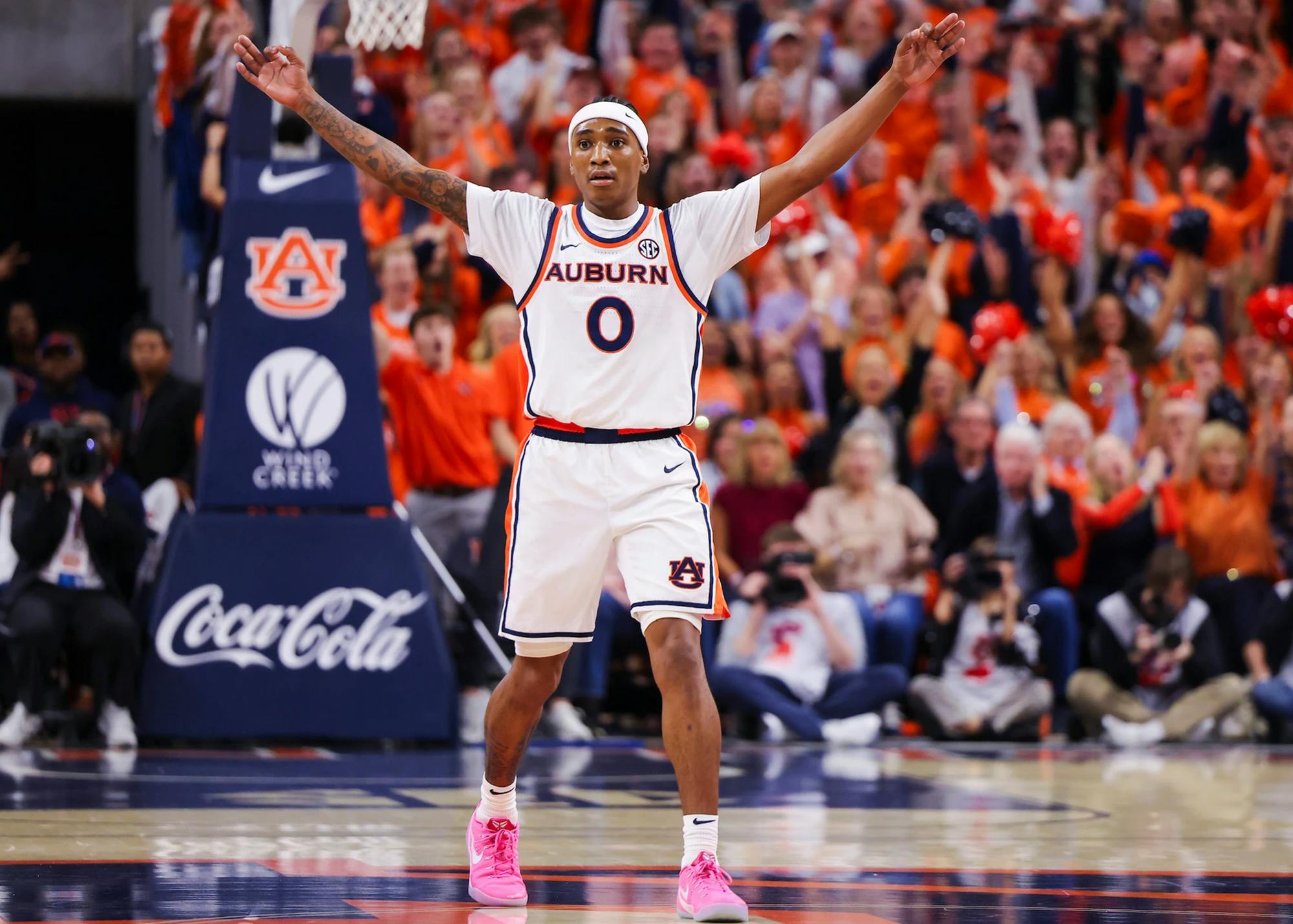 An Auburn men’s basketball player wearing a white jersey with number 0 raises both arms on the court, signaling to teammates or the crowd. The player wears bright pink shoes, and a packed arena of fans in orange cheers in the background.