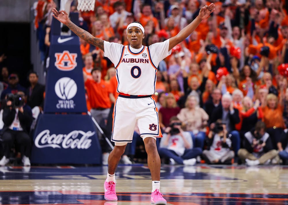 An Auburn men’s basketball player wearing a white jersey with number 0 raises both arms on the court, signaling to teammates or the crowd. The player wears bright pink shoes, and a packed arena of fans in orange cheers in the background.
