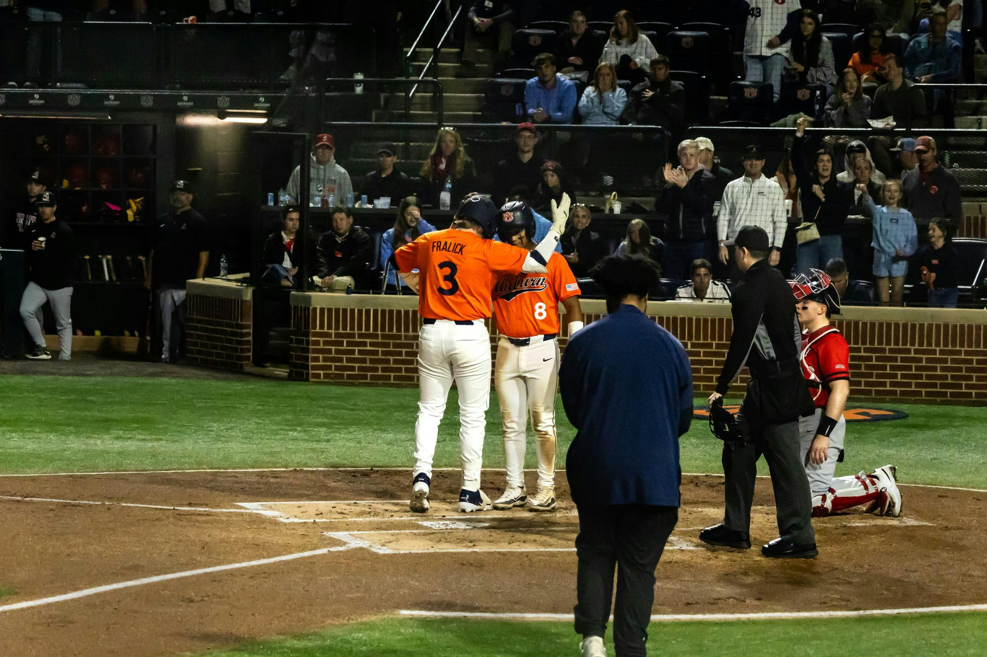 Two Auburn baseball players in orange jerseys celebrate at home plate during a night game, with one raising his arm as they meet. A catcher in red gear kneels nearby and an umpire stands behind the plate, while fans in the stands watch and applaud in the background.