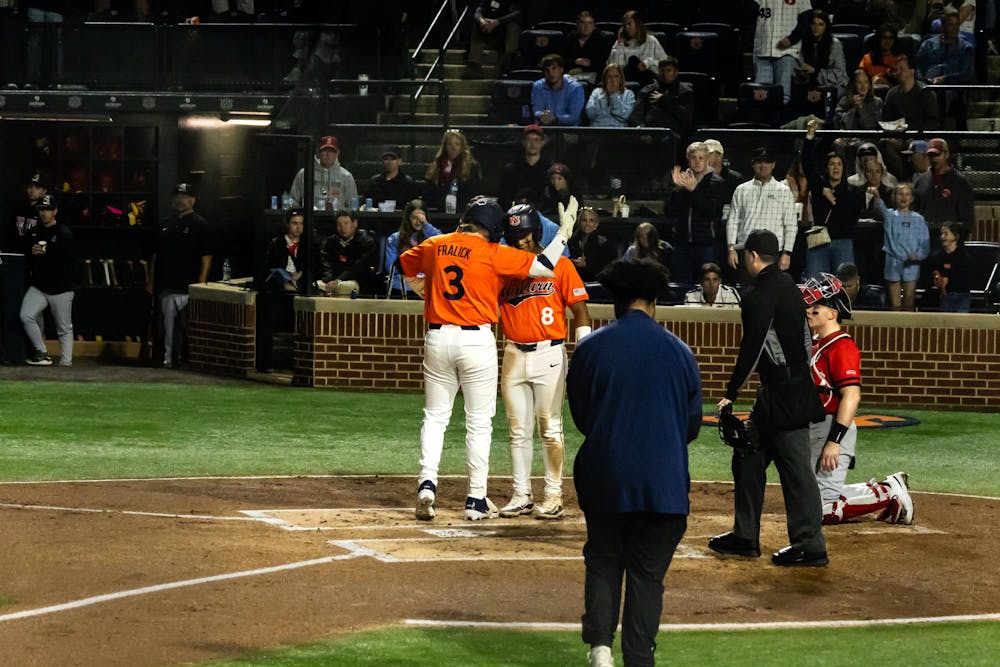 Two Auburn baseball players in orange jerseys celebrate at home plate during a night game, with one raising his arm as they meet. A catcher in red gear kneels nearby and an umpire stands behind the plate, while fans in the stands watch and applaud in the background.