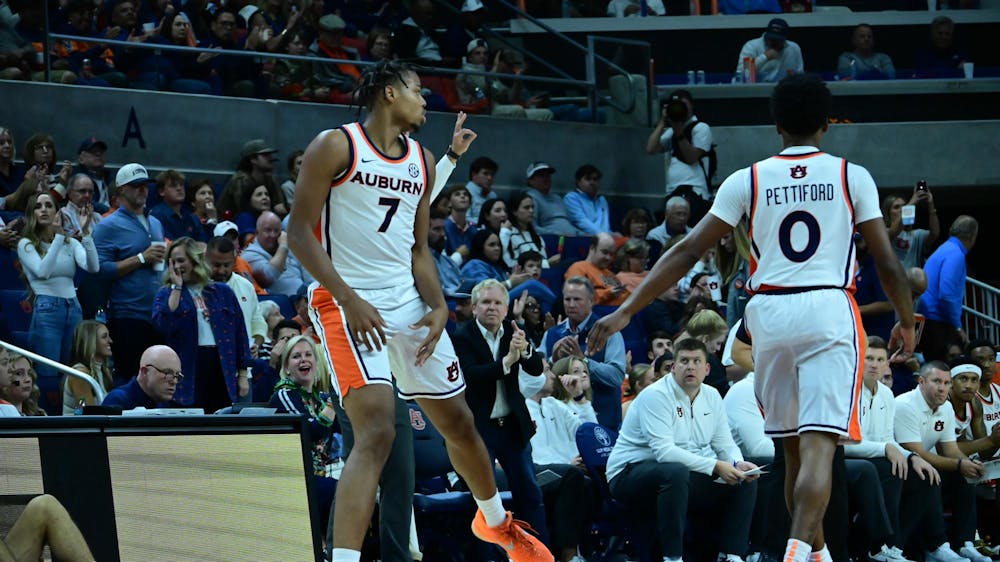 Two Auburn basketball players celebrate during a game, one wearing jersey number 7 and the other number 0. The player facing the camera gestures energetically while the other turns toward him. Coaches, teammates, and fans in the stands watch and applaud from the background in a lively arena setting.