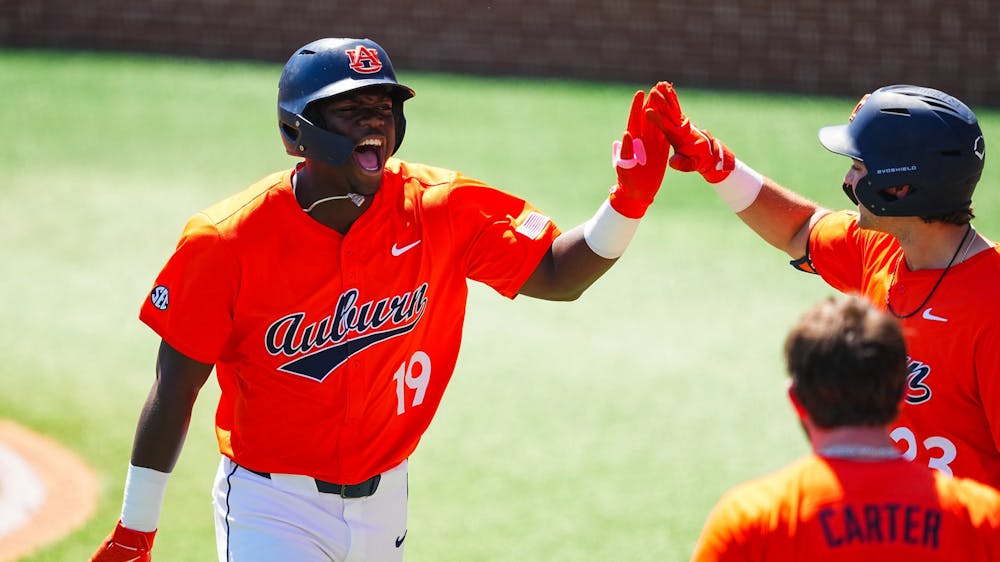 A baseball player in an orange Auburn jersey (number 19) celebrates energetically, shouting with excitement as he high-fives a teammate near the dugout. Another teammate with “Carter” and number 5 on his back stands in the foreground, while the green field and infield dirt are visible in the background.