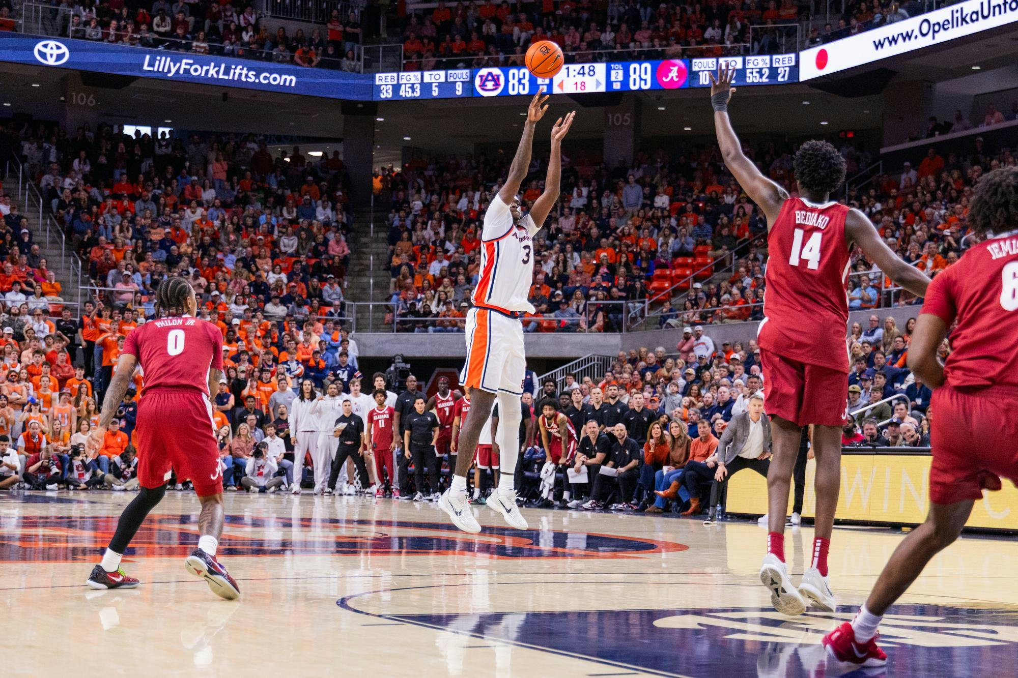 A basketball player in a white uniform jumps to shoot while defenders in red uniforms position themselves nearby, and a crowded arena watches.