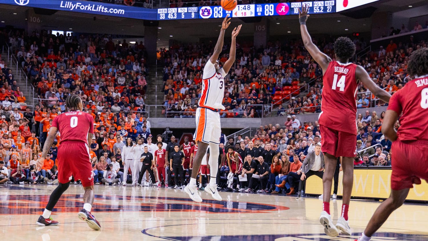 A basketball player in a white uniform jumps to shoot while defenders in red uniforms position themselves nearby, and a crowded arena watches.