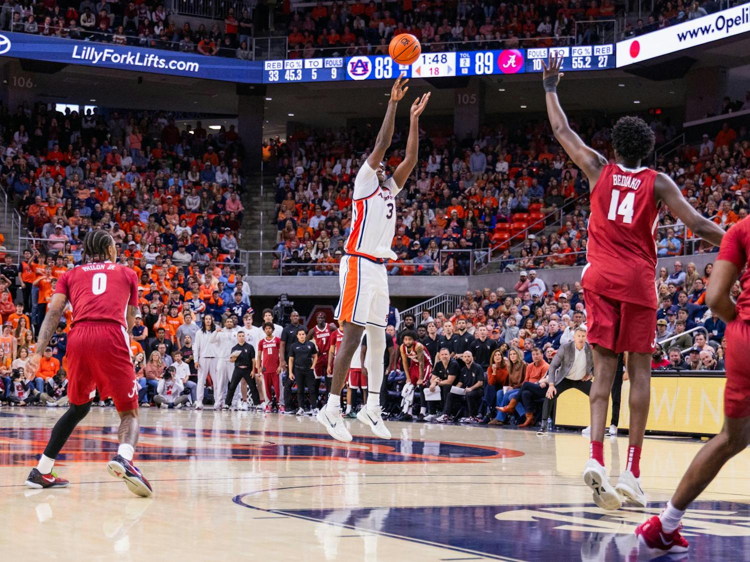 A basketball player in a white uniform jumps to shoot while defenders in red uniforms position themselves nearby, and a crowded arena watches.