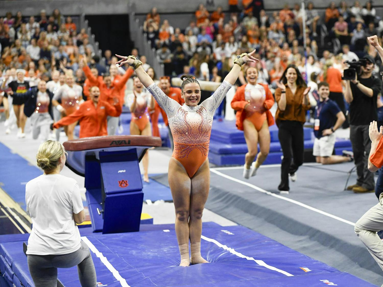 A gymnast celebrates with outstretched arms in a vibrant arena while teammates and spectators cheer enthusiastically.