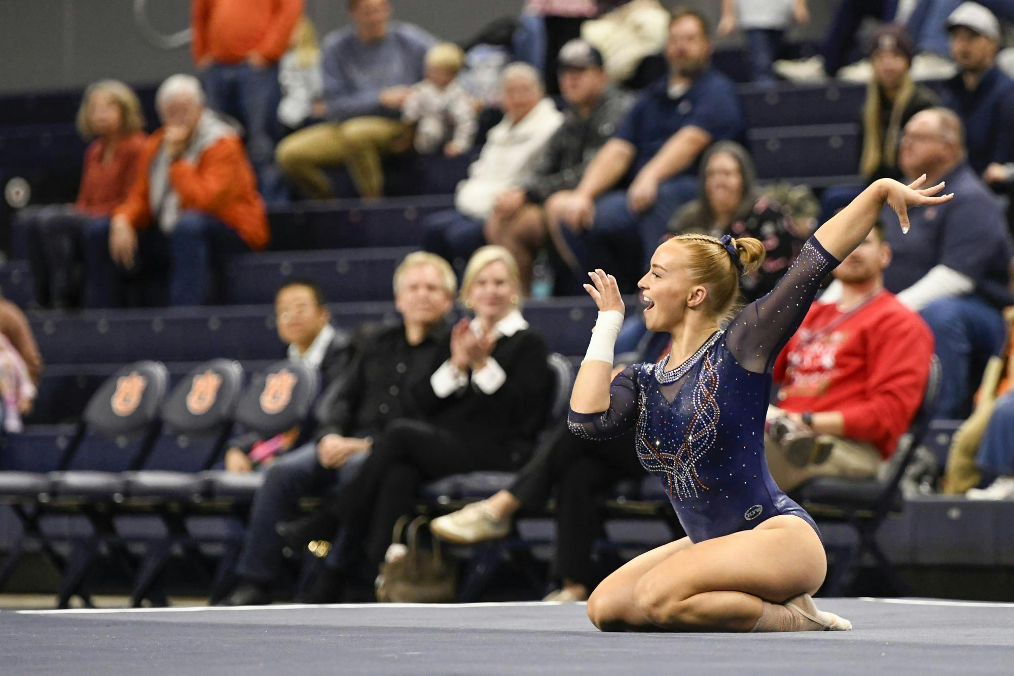 A gymnast in a sparkly blue leotard is kneeling on the mat, performing a gesture, while a captivated audience watches from the stands.