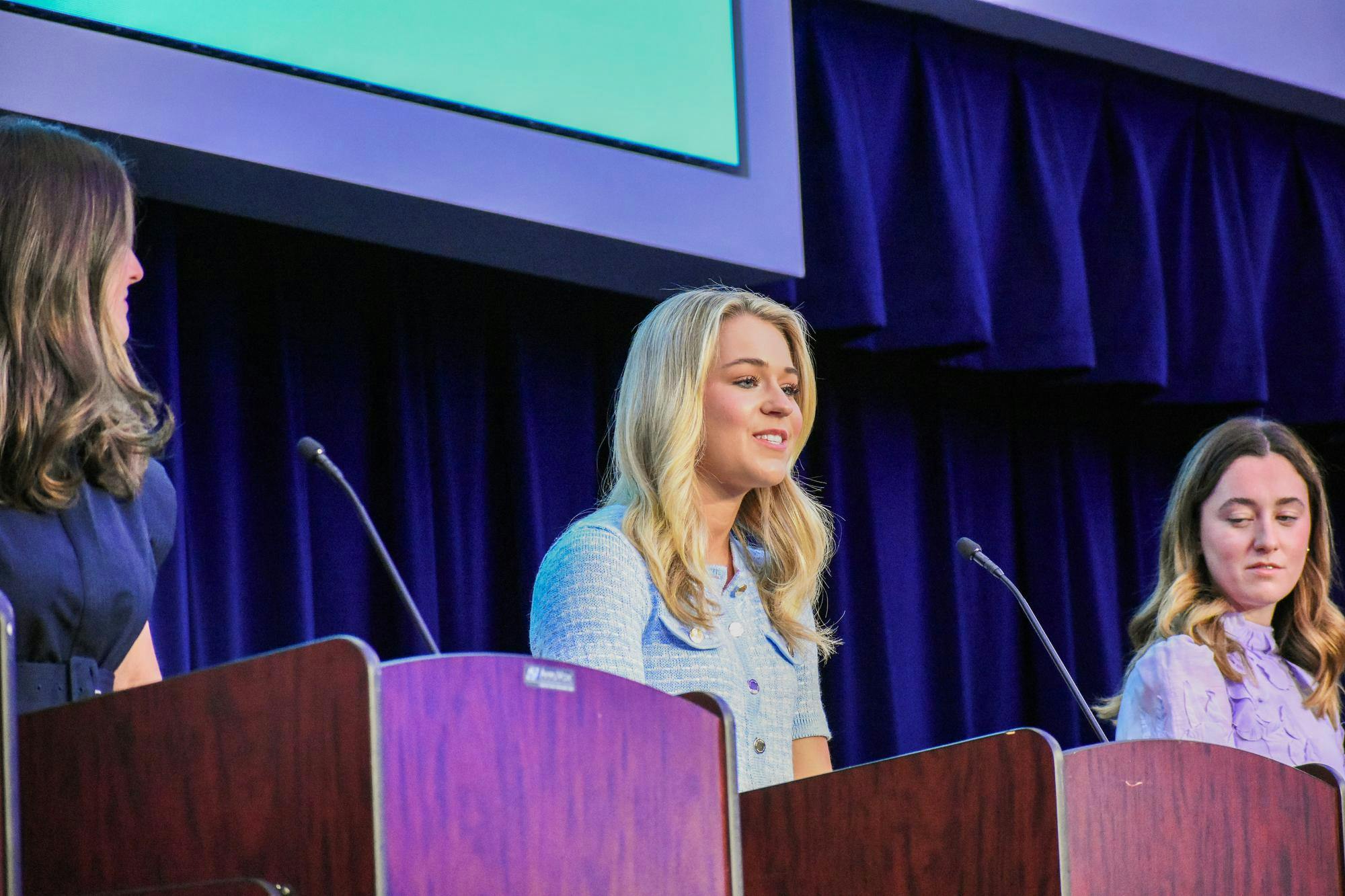 Three women are standing behind podiums, with one speaking while the others listen attentively.