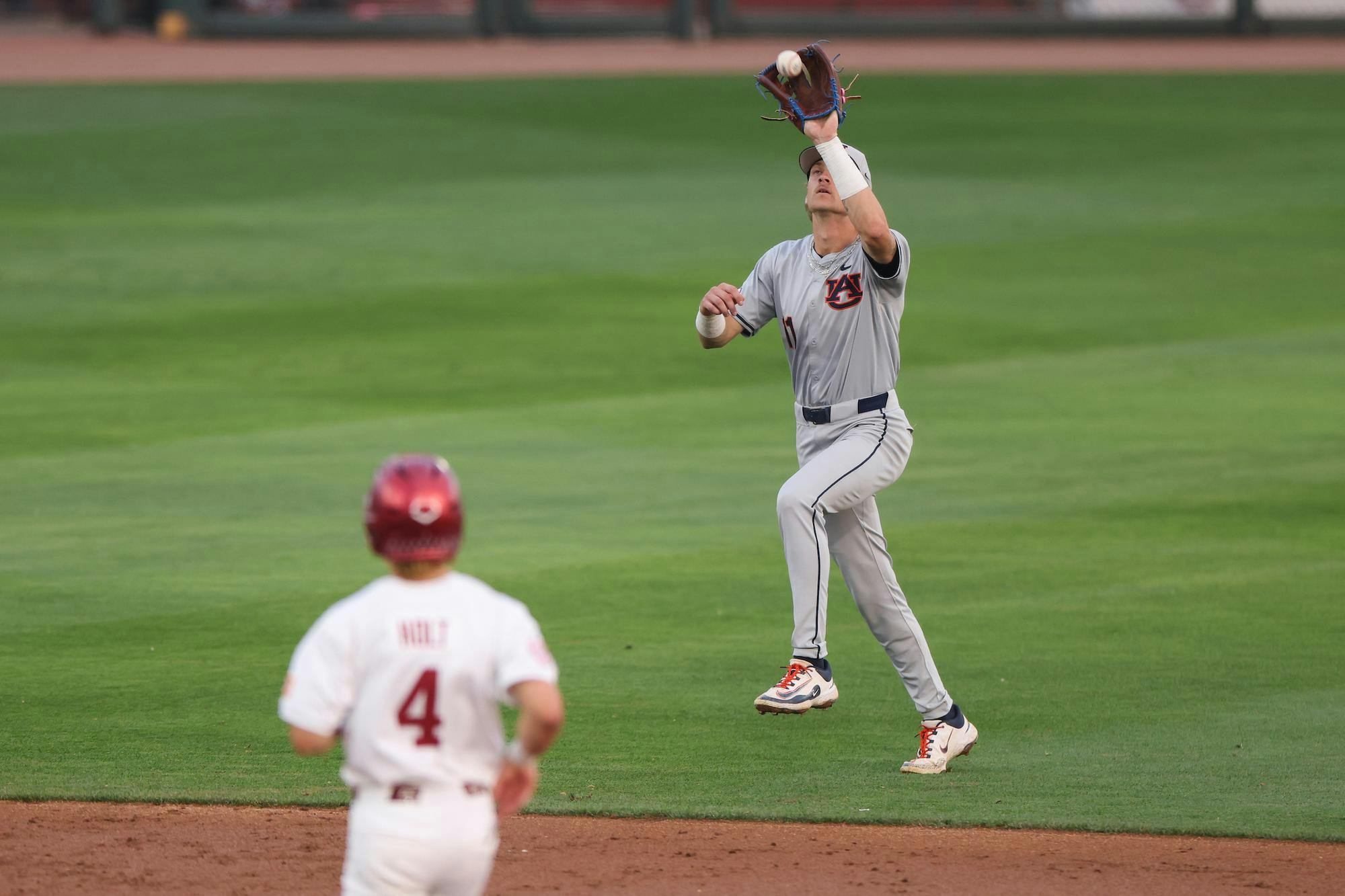 A player in gray baseball attire jumps to catch a ball while another player in red watches from a distance.