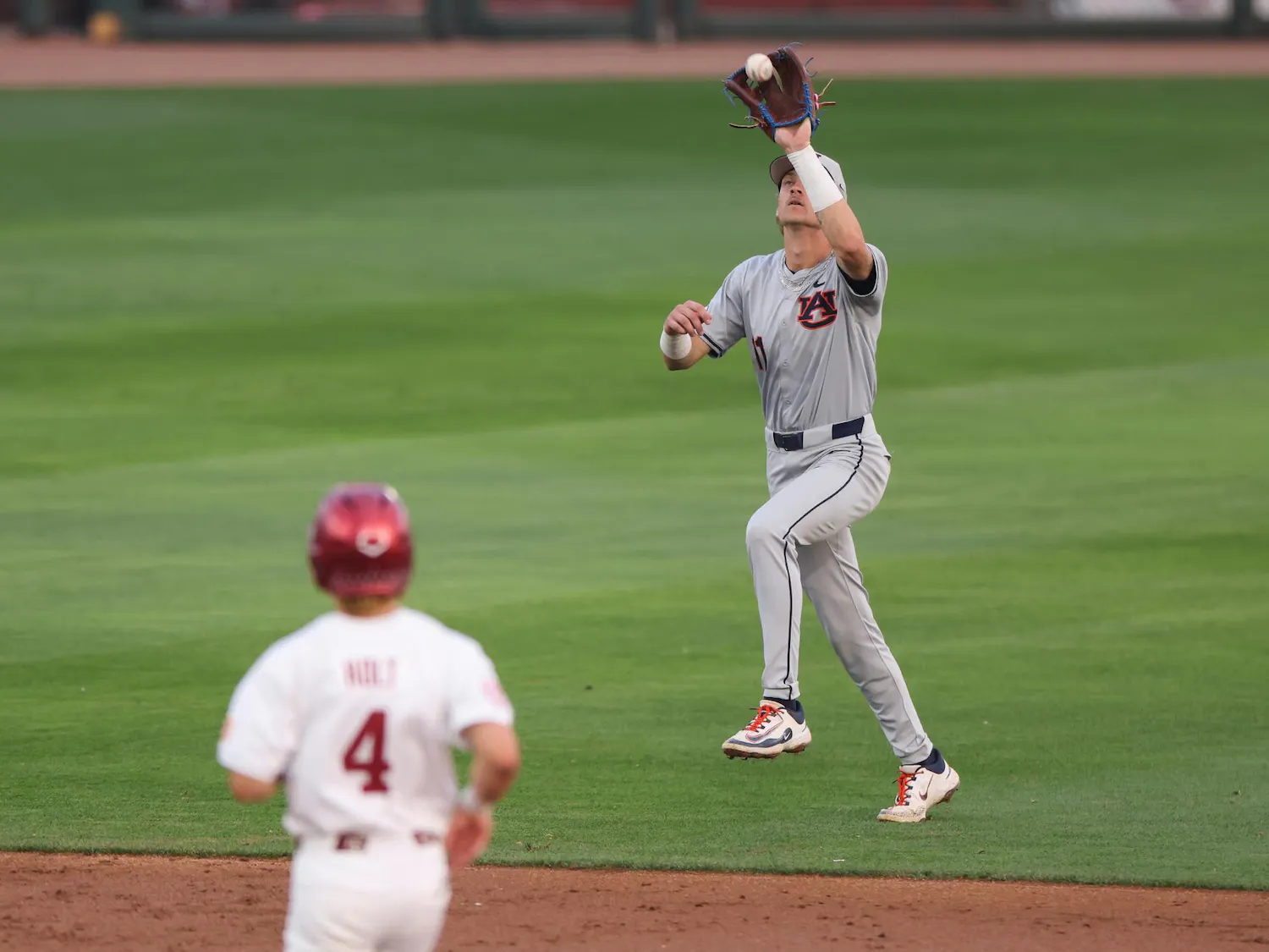 A player in gray baseball attire jumps to catch a ball while another player in red watches from a distance.